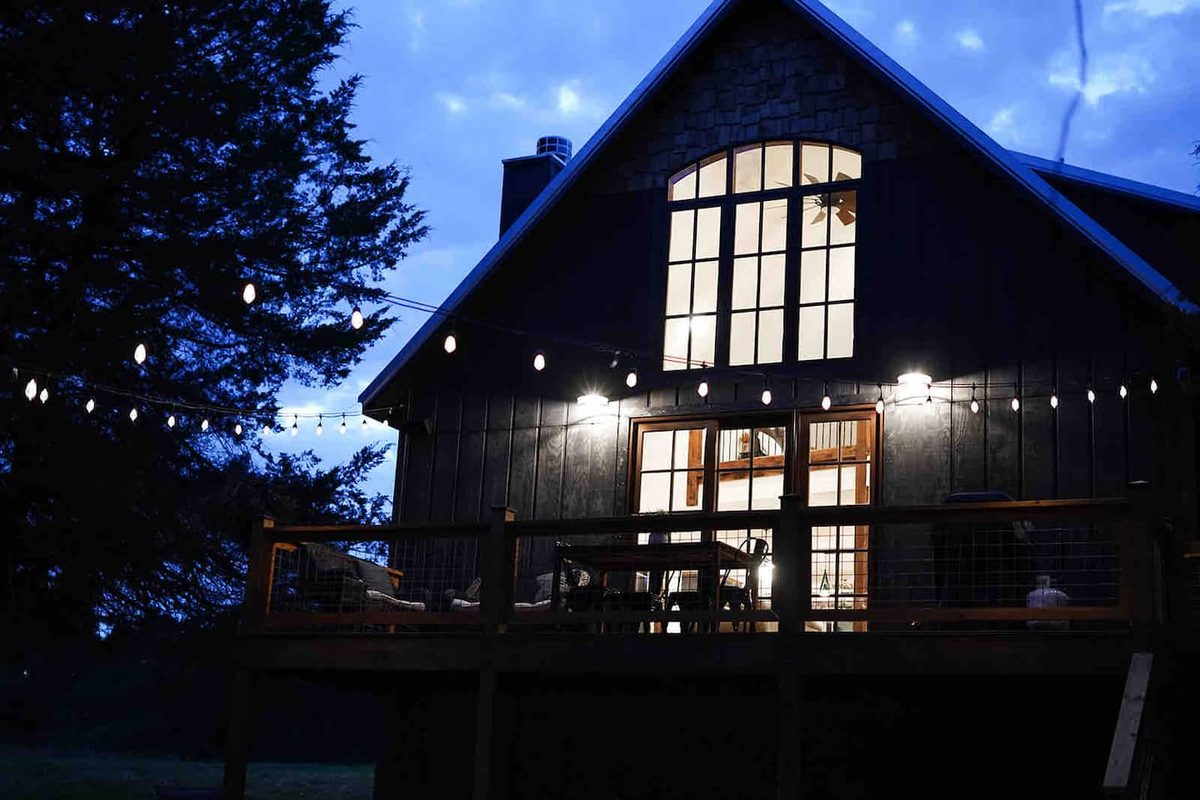 The cabin glowing at dusk with warm light through its tall windows and wraparound deck